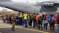 Zahlreiche Personen stehen in einer Schlange vor einem Flugzeug. Davor steht ein bewaffneter Soldat.