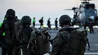 Mehrere Soldaten in Flecktarnuniform stehen hintereinander auf dem Flugdeck eines Schiffs; im Hintergrund ein Hubschrauber.