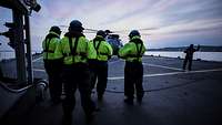 Auf dem Flugdeck eines Schiffs steht ein Hubschrauber, davor mehrere Soldaten in blau-gelber Arbeitskleidung.