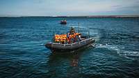 Ein Festrumpfschlauchboot besetzt mit mehreren Menschen die orangene Westen tragen fährt übers Wasser