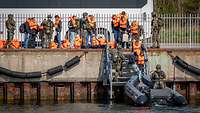 Personen mit Schwimmwesten und Soldaten stehen auf einer Hafenmauer. Im Vordergrund ein Speedboot. 