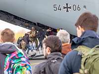 Personen stehen in einer Schlange vor einem Flugzeug. Ein Soldat hilft beim Einsteigen.