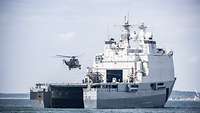 Ein graues Schiff in See; hinten ein flaches Flugdeck mit zwei Hubschraubern.