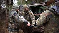 Drei Soldaten stehen neben einem Fahrzeug im Wald, zwei halten eine Mörserpatrone