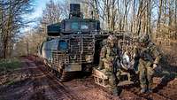 Auf einem Waldweg steht ein Panzer. An seinem Heck stehen mehrere Soldaten.