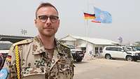 Portrait eines Soldaten im Feldanzug mit Wüstentarn. Er steht im Freien, hinter ihm wehen die deutsche und die UN-Flagge.
