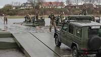 Ein Geländewagen vom Typ Wolf fährt über eine Rampe auf eine Schwimmbrücke mit deutschen und britischen Soldaten.