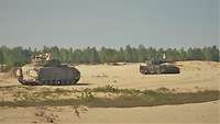 Zwei Schützenpanzer stehen auf sandigem Boden, im Hintergrund Bäume.