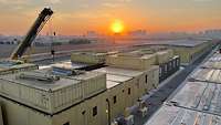 Mehrere Container stehen nebeneinander und im linken Bildrand steht ein Kran. Im Hintergrund geht die Sonne über Erbil auf.