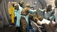 Mehrere Soldaten in Schutzanzügen verpflegen im Zuge einer Übung einen verwundeten Soldaten in einem aufblasbaren Zelt.