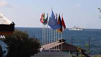 Eine Reihe von Masten mit den Flaggen der am Einsatz beteiligten Nationen, dahinter im Meer ein UN-Schiff