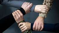 Fünf verschiedene Arme greifen sich gegenseitig am Handgelenk und bilden so eine Einheit. Jeder Arm trägt eine andere Uniform. 
