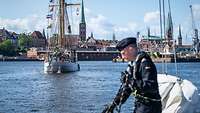 Ein kleines Segelschiff in einem Hafen vor einer Stadtkulisse mit mehreren Kirchtürmen.