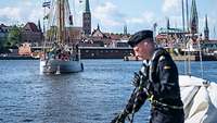 Ein kleines Segelschiff in einem Hafen vor einer Stadtkulisse mit mehreren Kirchtürmen.