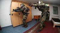 Zwei Soldaten mit Helm und Schutzweste stehen mit ihren Gewehren im Anschlag in einem Zimmer.