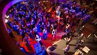 Viele Menschen tanzen vor einer Bühne auf dem Parkett in einem blau-rot erleuchteten Saal.