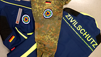 Eine Bundeswehr Uniform und eine Jack vom Zivilschutz die übereinander liegen beide mit dem Zeichen des BBK