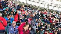 Viele Menschen sitzen in einem Block vom Stadion und schwenken Fanschals