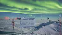 Ein Schild mit Informationen zu Roald Amundsen und Robert F. Scott steht in einer Eiswüste. Am Himmel sind Polarlichter.