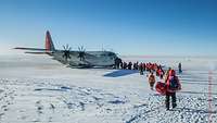 Auf einer Schneefläche steht ein Transportflugzeug. Eine Gruppe von Personen in dicker, roter Schneeschutzkleidung steigt ein.