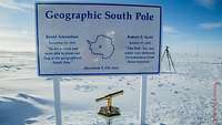 In einem Eismeer steht ein Schild mit der Aufschrift „Geographic South Pole“. Davor ist ein spezieller Marker angebracht.