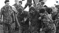 Schwarzweiß-Aufnahme von 1982. Britische Soldaten helfen einem verwundeten Kameraden aus einem Landungsboot an Land