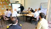 A group of participants is sitting at tables while a soldier is explaining the situation on the interactive whiteboard.