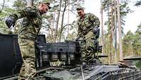Zwei Soldaten stehen mit einem Bauteil in der Hand auf einem Panzer im Wald.