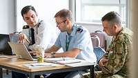 Three soldiers from different nations are discussing something on the laptop in front of them.