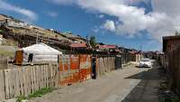 Blick entlang einer unbefestigten Dorfstraße, an einem Hang stehen Häuser und eine Jurte.