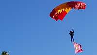 Ein Fallschirmspringer der spanischen Armee schwenkt im Sinkflug vor strahlend blauem Himmel die Spanische Flagge.