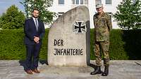 Ein Zivilist im dunklen Anzug und ein Soldat stehen links und rechts neben dem Stein der Infanterie.