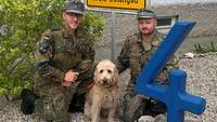 Zwei Soldaten knien vor einem Ortsschild. In ihrer Mitte sitzt ein Labradudel mit hellem Fell.