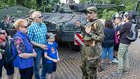 Ein Soldat steht neben einer Besuchergruppe und spricht. Hinter ihm sind weitere Besucher und ein Panzer zu sehen.