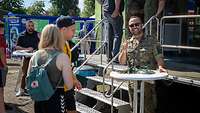 Ein Soldat steht an einem weißen runden Tisch vor dem Karrieretruck. Neben ihm stehen zwei Besucher vom Tag der Bundeswehr.