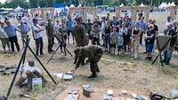 Ein Soldat steht auf einer Wiese und zeigt Zuschauern verschiedene Gegenstände.