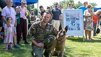 Ein Soldat kniet neben einem Schäferhund. Zuschauer sind im Hintergrund zu sehen.