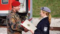 Ein Oberst in Bundeswehr-Uniform überreicht einer Feuerwehrfrau auf einem großen sonnigen Platz eine Urkunde