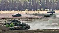 Fünf Panzer fahren mit hoher Geschwindigkeit über ein weites braunes Feld in der Sommerhitze.