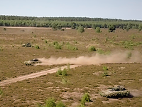 Mehrere Panzer fahren mit hoher Geschwindigkeit über ein weites braunes Feld in der Sommerhitze.