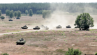 Zahlreiche Panzer fahren mit hoher Geschwindigkeit über ein weites braunes Feld. Staub wirbelt auf.