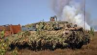 Ein Panzer steht vor einem braunen Panzerwrack und einer Rauchwolke auf einer Heide im Sommerlicht.