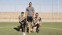 Drei Personen und ein Hund befinden sich auf einem Sportplatz und schauen in die Kamera