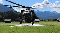 Ein Hubschrauber vom Typ CH-53 landet auf einem Helipad auf einer Wiese. Im Hintergrund sind Berge zu sehen.