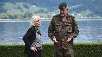 Eine grauhaarige Frau steht mit einem Soldaten vor einem See. Beide unterhalten sich.