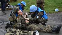 Ein Soldat liegt verletzt am Boden, zwei Soldaten mit blauen Helmen stützen und untersuchen ihn. 