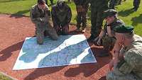 Six soldiers have spread out around a map and are looking at it. They are in the open air.