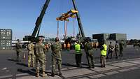 Soldatinnen und Soldaten der NATO stehen an einem Binnenhafen und sehen, wie ein LKW mit zwei Kränen vom Schiff gehoben wird.