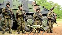 Vier bewaffnete Infanteristen stehen vor einem Gefechtsfahrzeug, vor ihnen ein Roboterhund.