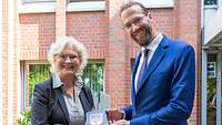 Eine Frau und ein Mann in schmuckem Zivil stehen vor einem großen Haus und halten ein Wappen in der Hand